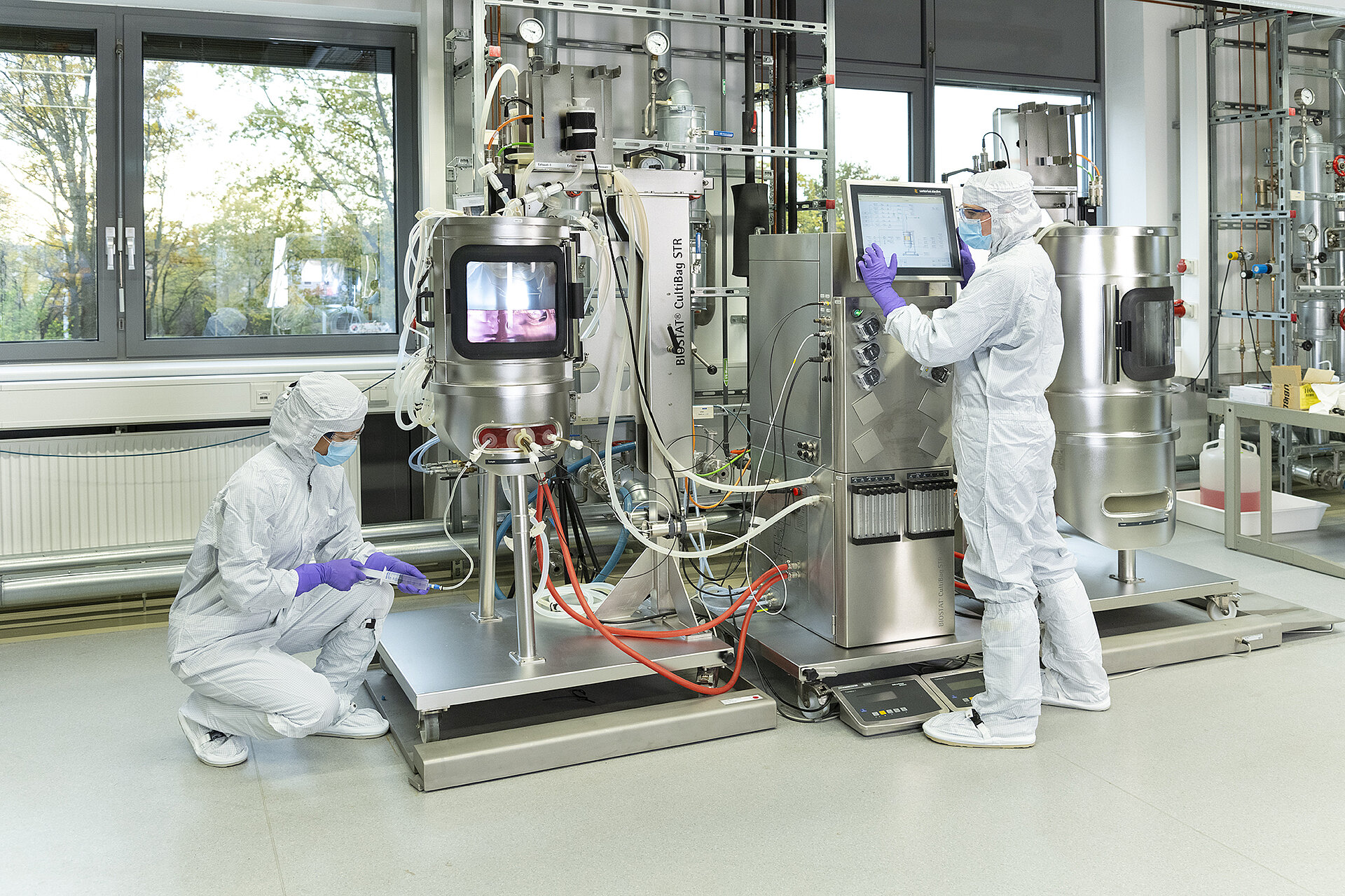 Zwei Personen in Schutzkleidung bedienen und überwachen eine Bioprozessanlage mit Edelstahlbehältern und Schläuchen in einem Labor.